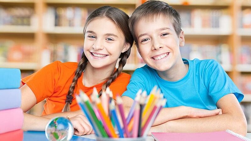 Lachende Schülerin und Schüler im Klassenzimmer vor Stiften und Arbeitsmaterialien