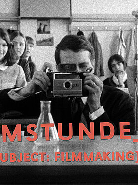 Schwarzweißaufnahme: Hintgrund Schülerinnen und Schüler im Klassenzimmer, Vordergrund kniender Fotograf mit einer alten Polaroid-Kamera. Text in rot: FILMSTUNDE_23 [Subject: Filmmaking]