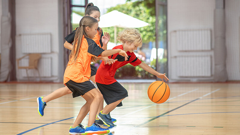 Kinder in heller Sportbekleidung spielen zusammen Basketball in Turnhalle. 