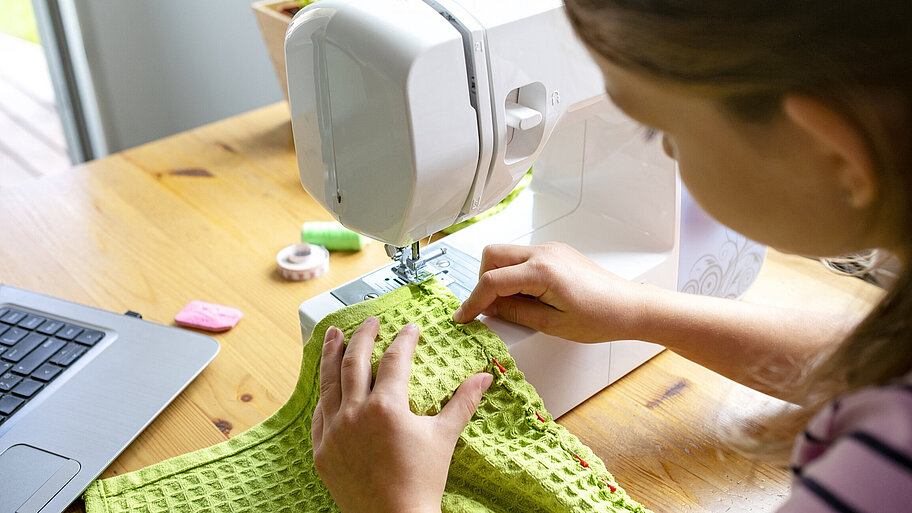 Mädchen arbeitet an Nähmaschine und lernt mit Laptop online zu nähen