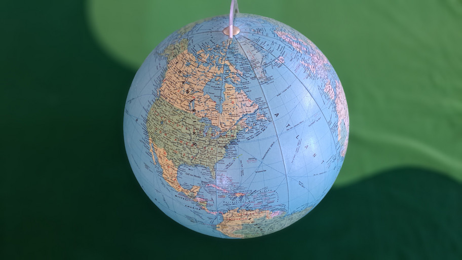 Auf dem Bild ist ein Globus zu sehen, der die Erde darstellt. Der Globus hängt an einer Schnur und zeigt vor allem den amerikanischen Kontinent, einschließlich Nord- und Südamerika. Der Hintergrund ist grün, vermutlich eine Wand oder ein Vorhang. Der Globus selbst ist detailliert mit geografischen Merkmalen und Ländergrenzen versehen.