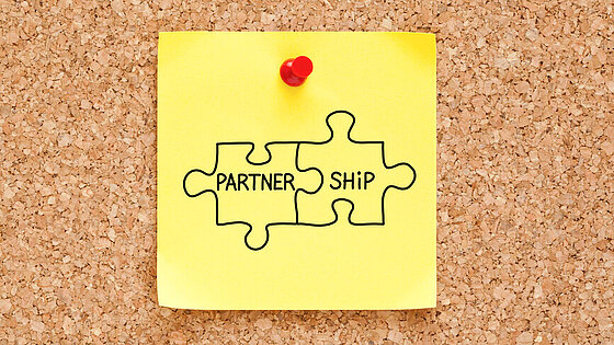 Korkwand mit einem gelben Postit auf dem zwei Puzzleteile zu sehen sind mit den Partner Ship