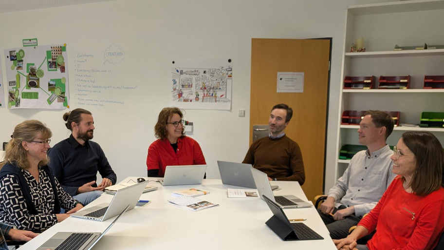 Fotodarstellung von sieben Menschen, die um einen Tisch in einer Besprechung sitzen