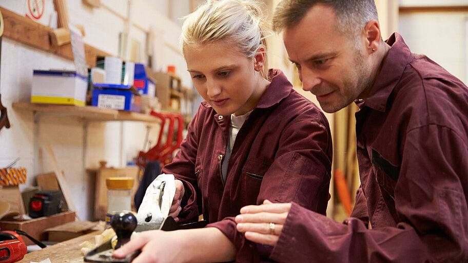 Carpenter With Apprentice Planing Wood In Workshop