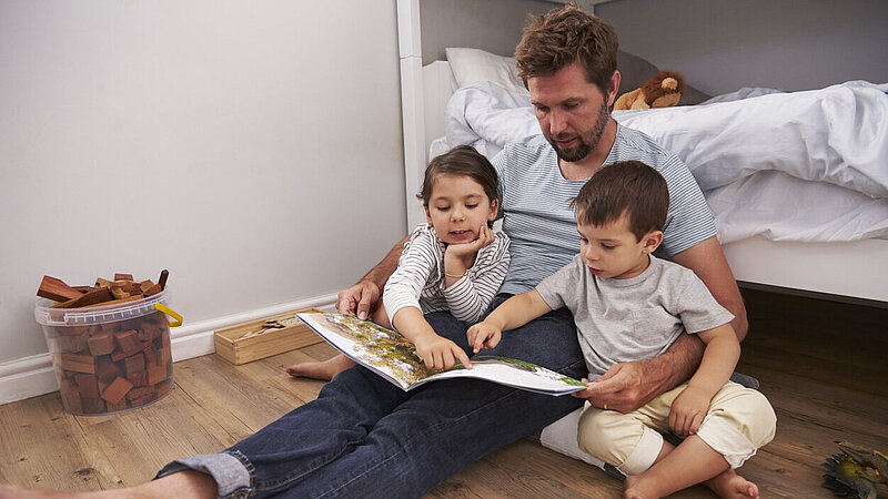 Vater und Kinder sitzen im Kinderzimmer am Boden und lesen gemeinsam ein Buch