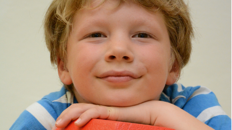 Ein Junge hält einen großen, orangefarbenen Würfel mit schwarzen Zahlen in den Händen. Die sichtbaren Zahlen auf dem Würfel sind 3 und 4.