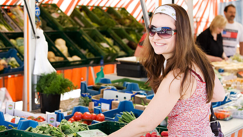 Lächelnde junge Frau mit Sonnenbrille steht am Gemüsestand