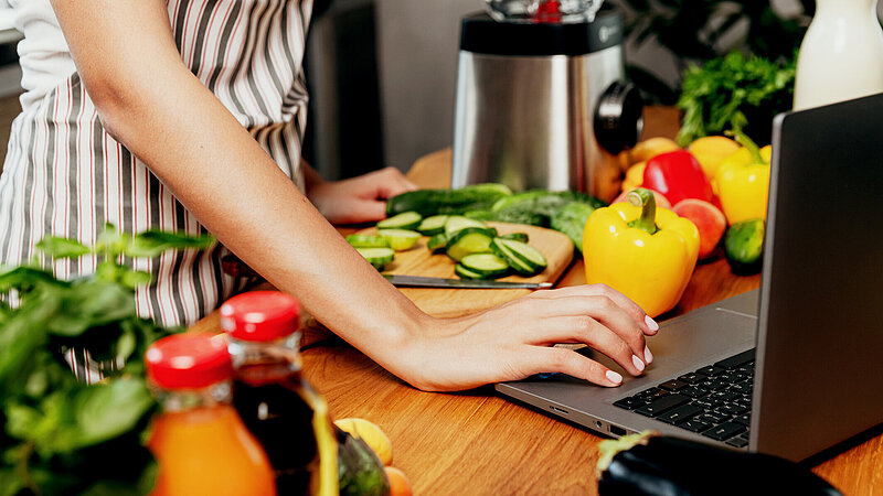 Weibliche Hand mit Laptop in der Küche beim Gemüseschneiden