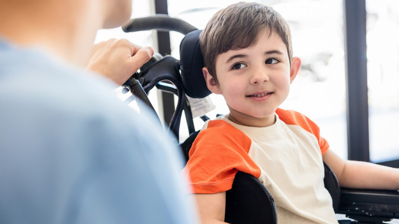 Beautiful little boy on wheelchair smiling and looking with admiration at his unrecongizable physical therapist