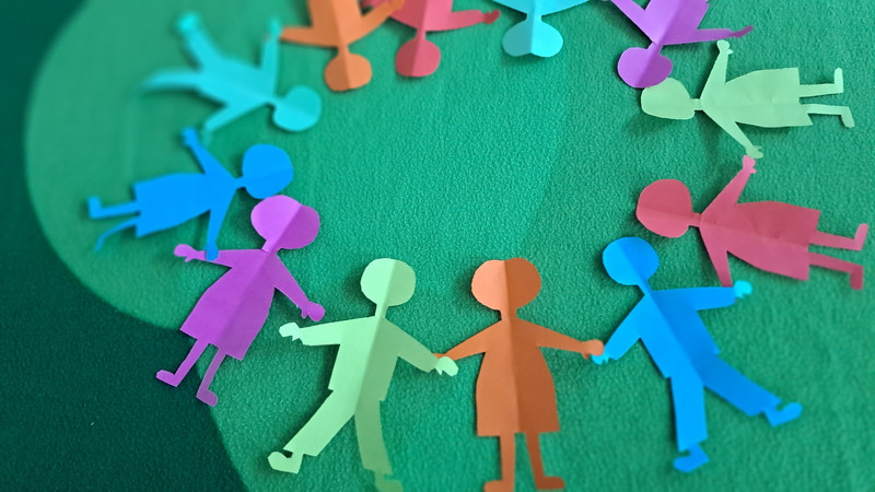 Auf dem Bild sind bunte, ausgeschnittene Papierfiguren von Kindern zu sehen, die im Kreis stehen und sich an den Händen halten. 