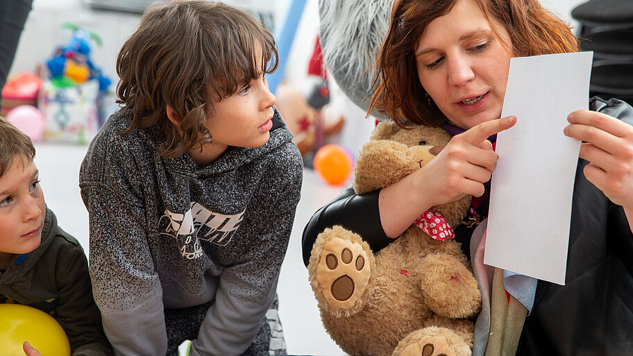 Lehrerin mit Teddy im Arm zeigt Schülern ein weißes Papier