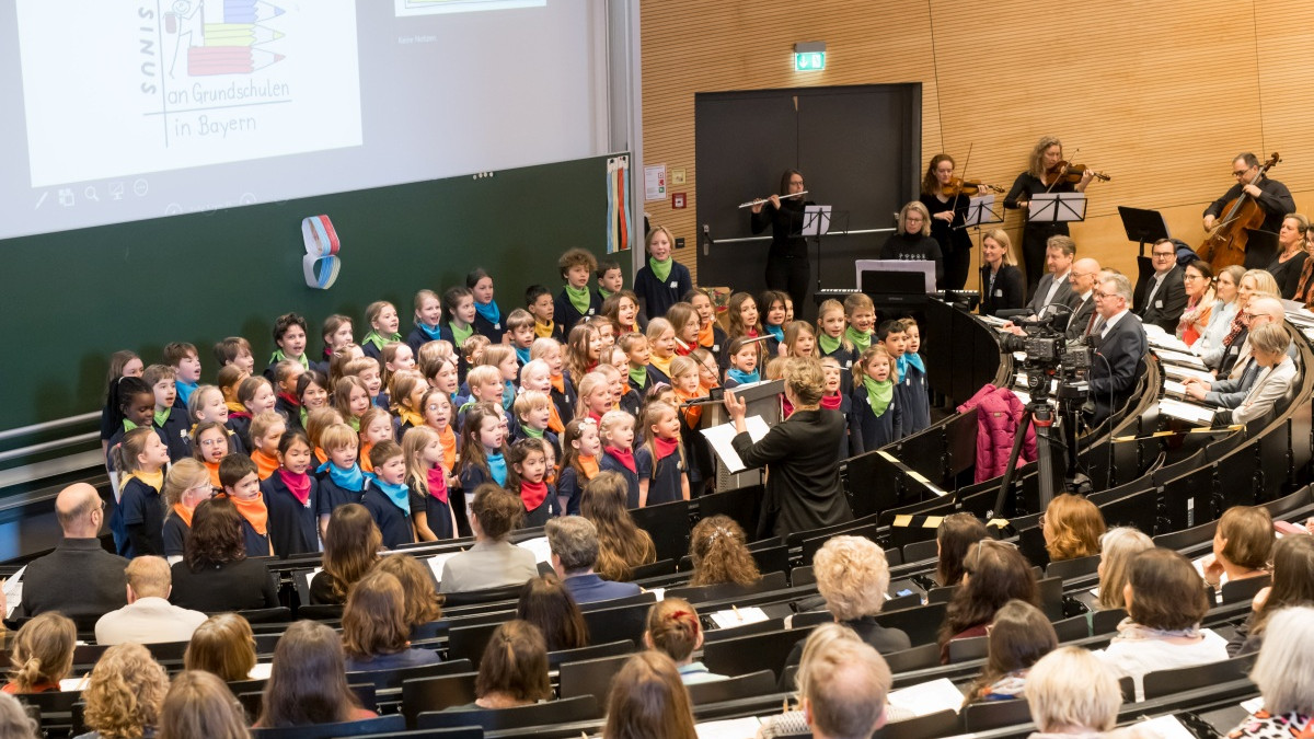 Zu sehen ist ein großer Hörsaal mit dem feierlichen Auftakt der Veranstaltung SINUS goes QuaMath. Vorne steht ein Chor aus Kindern, die bunte Halstücher tragen und singen. Eine Dirigentin leitet den Chor. Im Hintergrund spielt ein kleines Orchester mit verschiedenen Instrumenten. Im Publikum sitzen zahlreiche Erwachsene, die aufmerksam der Aufführung folgen. An der Wand hinter dem Chor ist eine große Tafel zu sehen, auf welche das SINUS-Logo projiziert wird.