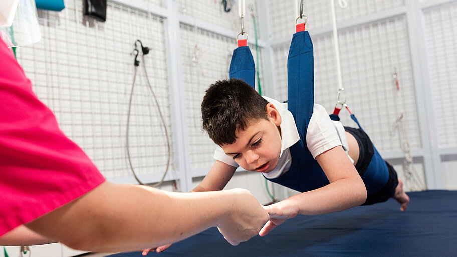 Junge mit Behinderung hängt während einer physiotherapeutischen Übung in einer Therapieschaukel